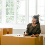 Content lady in casual clothes writing with pen on cardboard box while sitting near window in daylight in apartment preparing to relocate