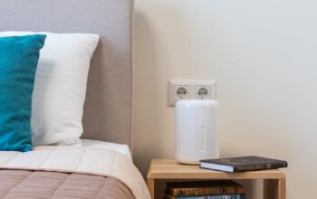 Stack of books and humidifier arranged on small minimalist wooden night table placed near comfortable bed with pillows in light room