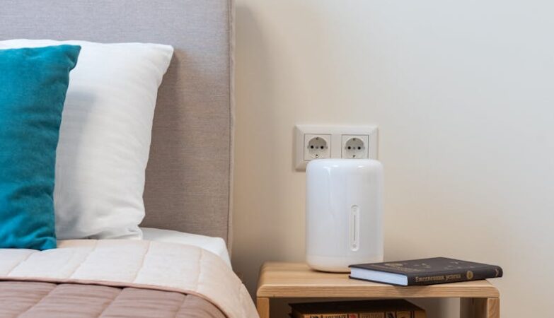 Stack of books and humidifier arranged on small minimalist wooden night table placed near comfortable bed with pillows in light room